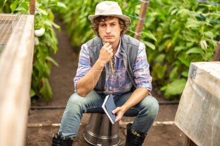 Photo for Serious pensive agriculturist with a pencil and notebook in his hands sitting on the overturned bucket in the hothouse - Royalty Free Image