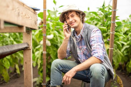 Photo for Smiling contented agriculturist sitting on the overturned bucket in the greenhouse during the phone call - Royalty Free Image