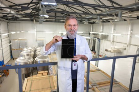 Foto de Pleased brewery technologist in a lab coat and eyeglasses holding a tablet computer in his hands - Imagen libre de derechos
