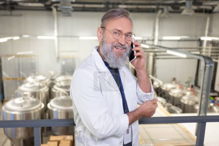 Foto de Waist-up portrait of a smiling contented brewery technologist in eyeglasses talking on the smartphone in the factory - Imagen libre de derechos