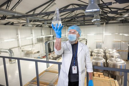 Téléchargez les photos : Serious concentrated brewery technologist in a protective mask examining the contents of a glass flask - en image libre de droit