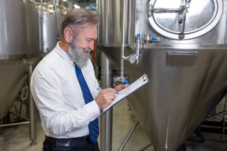 Téléchargez les photos : Focused brewery technologist in an elegant tie writing something with a ball-point pen on the clipboard - en image libre de droit
