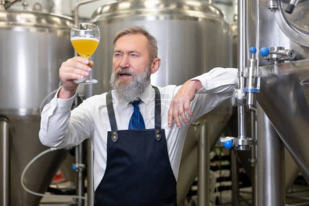 Téléchargez les photos : Waist-up portrait of a brewer leaning on the fermentation tank and looking at a glass of beer in his hand - en image libre de droit