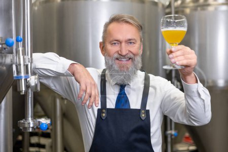 Foto de Waist-up portrait of a cheerful brewer leaning on the fermentation tank with a glass of fresh beer - Imagen libre de derechos