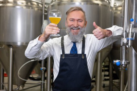 Foto de Joyful brewer in the apron leaning on the metal tank with a glass of drink and making a thumbs-up sign - Imagen libre de derechos