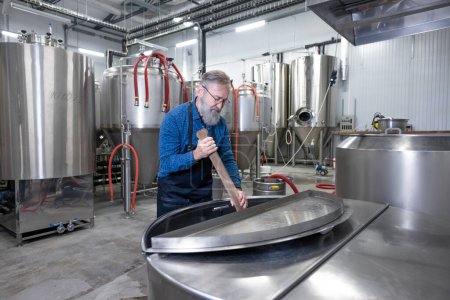 Téléchargez les photos : Stirring beer. Mature bearded factory worker stirring beer in a vat - en image libre de droit