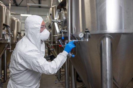 Foto de Experienced beer tech in a protective mask and gloves turning the manual pressure relief valve lever on the fermentation tank - Imagen libre de derechos