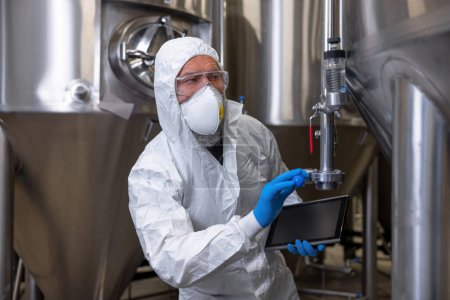 Foto de Waist-up portrait of a serious engineer with a tablet computer in the hand touching the knob of the pressure relief valve on the fermentation tank - Imagen libre de derechos