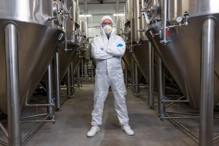 Téléchargez les photos : Full-length portrait of a serious brewery technologist in safety clothing standing between the metal tanks in the factory - en image libre de droit