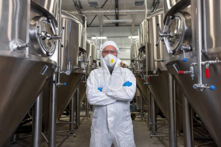 Foto de Technologist in a protective mask and nitrile gloves posing for the camera among the beer fermentation tanks - Imagen libre de derechos