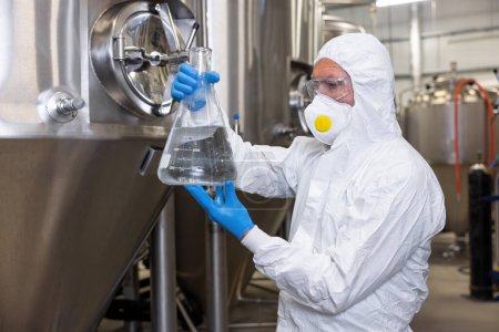 Téléchargez les photos : Serious focused brewery technologist staring at a glass flask with transparent liquid in his hands - en image libre de droit