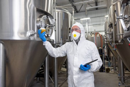 Foto de Beer maker in a protective mask and nitrile gloves holding a glass flask with a beverage and a tablet computer - Imagen libre de derechos