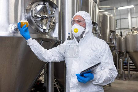 Foto de Serious brewer in nitrile gloves and a protective mask holding a glass flask with beer and a tablet computer - Imagen libre de derechos