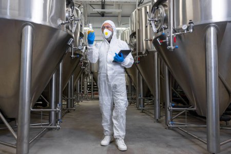 Téléchargez les photos : Full-size portrait of a focused brewer in a protective mask examining the color of beverage in the glass flask - en image libre de droit