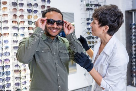 Foto de Optical store. Female optometrist helping a visitor to buy new eyeglasses - Imagen libre de derechos