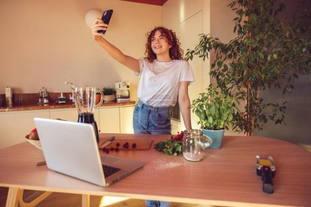 Téléchargez les photos : Selfie. Curly-haired young girl making selfie in the kitchen - en image libre de droit
