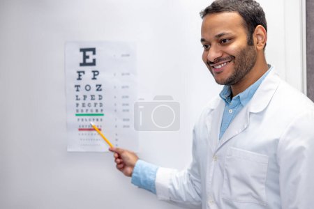 Photo for Ophtalmologist. Smiling male doctor standing near eyesight check list - Royalty Free Image