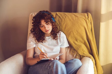 Foto de Diary. Curly-haired young girl in headphones sitting in the armchair and making notes in a diary - Imagen libre de derechos