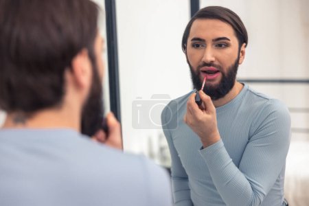 Foto de Calm focused transgender person applying the pink gloss to the lower lip before the mirror - Imagen libre de derechos