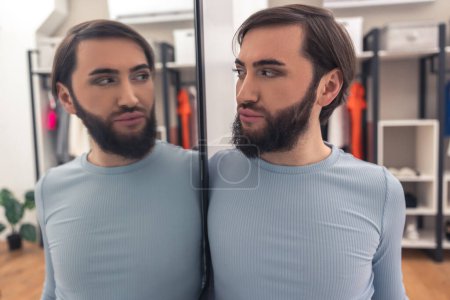 Téléchargez les photos : Serious young Caucasian transgender person looking at himself in the mirror in the fitting room - en image libre de droit
