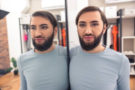 Téléchargez les photos : Portrait d'un homme barbu aux cheveux bruns avec un maquillage naturel sur le visage appuyé contre le miroir - en image libre de droit
