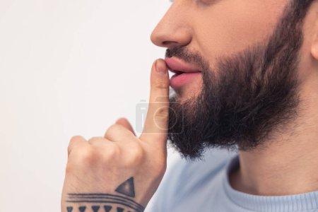Foto de Foto recortada de un hombre barbudo con el tatuaje de la mano presionando su dedo índice en la boca - Imagen libre de derechos