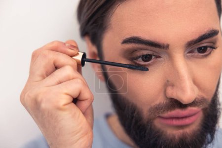 Photo for Closeup portrait of a serious pensive guy applying black mascara to his lower eyelashes with the wand - Royalty Free Image