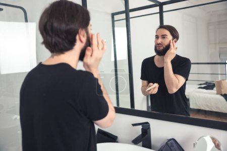 Téléchargez les photos : Focalisé jeune homme appliquant fond de teint crème sur la peau devant le miroir de salle de bain - en image libre de droit