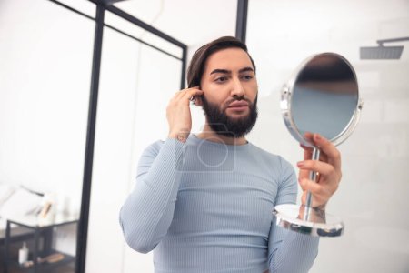 Photo for Waist-up portrait of a serious cute young man looking at his face in the hand-held mirror - Royalty Free Image
