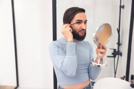 Photo for Focused guy applying black mascara to his long eyelashes while looking at the hand-held mirror - Royalty Free Image