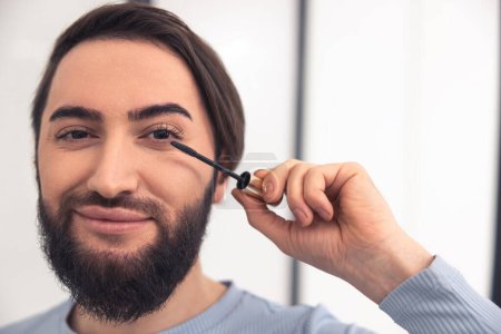 Photo for Headshot of a smiling contented man applying black mascara to his upper eyelashes using the wand - Royalty Free Image