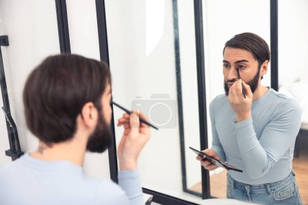 Photo for Serious focused dark-haired male applying the eyeshadows to the upper eyelid using a thin brush - Royalty Free Image