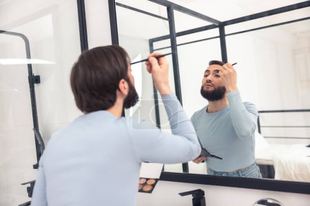 Photo for Serious concentrated young Caucasian man applying the eyeshadows to his upper eyelid with a thin brush - Royalty Free Image