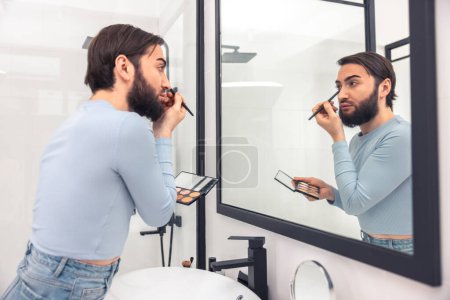 Téléchargez les photos : Focalisé homme caucasien appliquant les fards à paupières à sa paupière inférieure avant le miroir mural - en image libre de droit