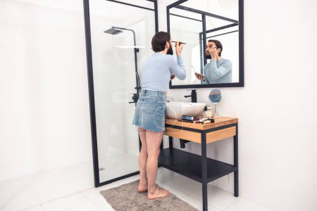 Téléchargez les photos : Homme dans la jupe denim tendance debout sur le tapis de sol tout en appliquant des cosmétiques devant le miroir de salle de bains - en image libre de droit