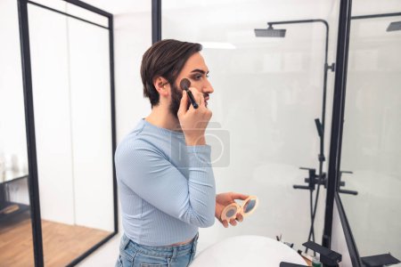 Photo for Side view of a cute guy applying compact powder to the cheeckbone before the mirror - Royalty Free Image
