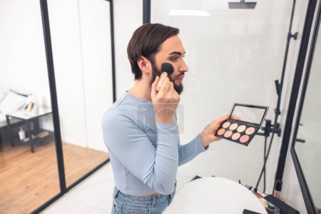 Photo for Side view of a serious guy applying blush to the cheekbone in front of the mirror - Royalty Free Image