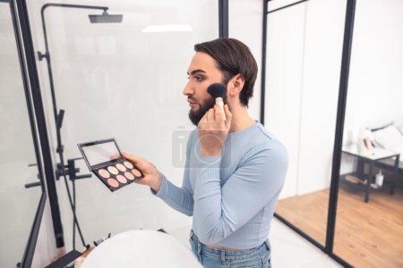 Photo for Waist-up portrait of a focused male holding a blusher box while applying rouge to the cheek - Royalty Free Image