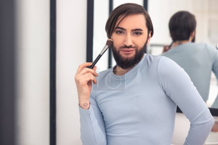 Photo for Waist-up portrait of a handsome guy standing at the wall-mounted mirror during the makeup application - Royalty Free Image