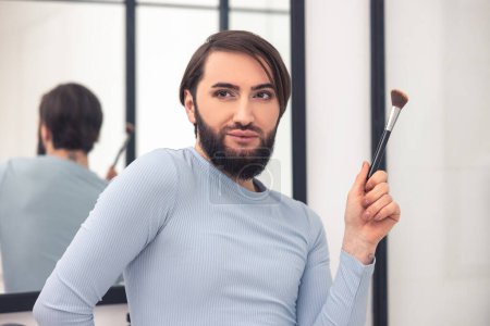 Téléchargez les photos : Portrait of a transgender person holding a cosmetic brush in the hand while standing at the bathroom mirror - en image libre de droit