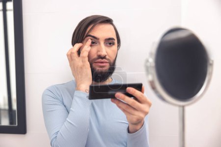 Téléchargez les photos : Portrait of a transgender person applying eyeshadows to the upper eyelid before the bathroom mirror - en image libre de droit