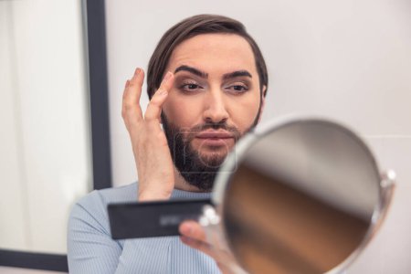 Téléchargez les photos : Headshot of a young man applying eyeshadows to the eyelids before the hand mirror in the bathroom - en image libre de droit