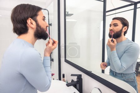Bearded dark-haired young man applying the pink lip gloss before the mirror in the bathroom