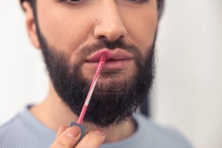 Photo for Cropped photo of a serious bearded mustached young Caucasian man applying the pink lip gloss - Royalty Free Image