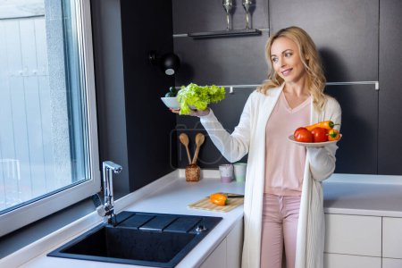 Téléchargez les photos : Joyeuse jeune femme appuyée contre le comptoir de la cuisine tout en tenant deux assiettes avec des légumes frais - en image libre de droit