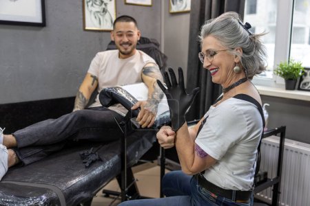 Téléchargez les photos : Tatoueuse. Mature maître de tatouage féminin travaillant avec le client - en image libre de droit