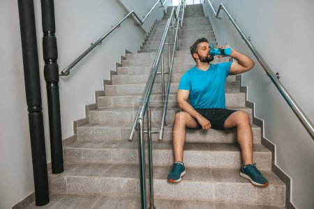 Photo for Thirsty after workout. Sportsman sitting on the stairs and drinking water - Royalty Free Image