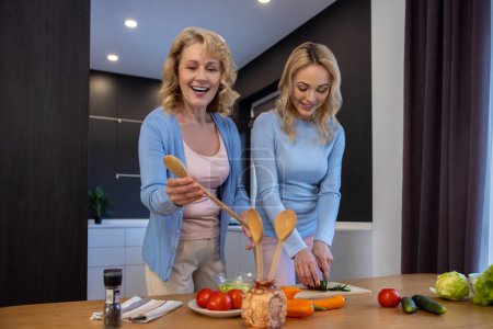 Photo for Joyous mature lady reaching for a wooden spoon while a young woman cutting fresh cucumbers - Royalty Free Image
