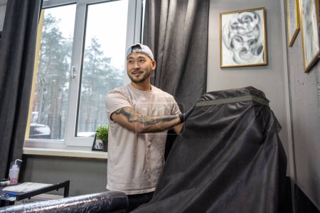 Photo for In the tattoo salon. Asian tattoo master preparing couch for tattoo session - Royalty Free Image