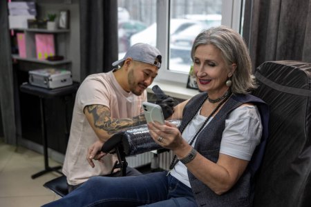 Téléchargez les photos : Dans le salon de tatouage. Femme aux cheveux gris parlant au téléphone pendant que le maître de tatouage travaille - en image libre de droit